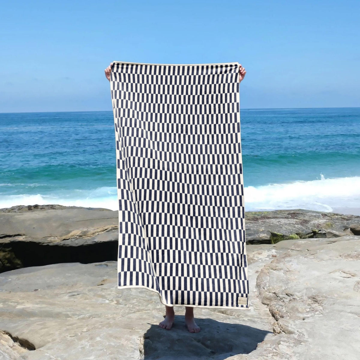 Person holding up the Shorebird Oversized Beach Towel in navy check on a rocky beach #color_nantucket