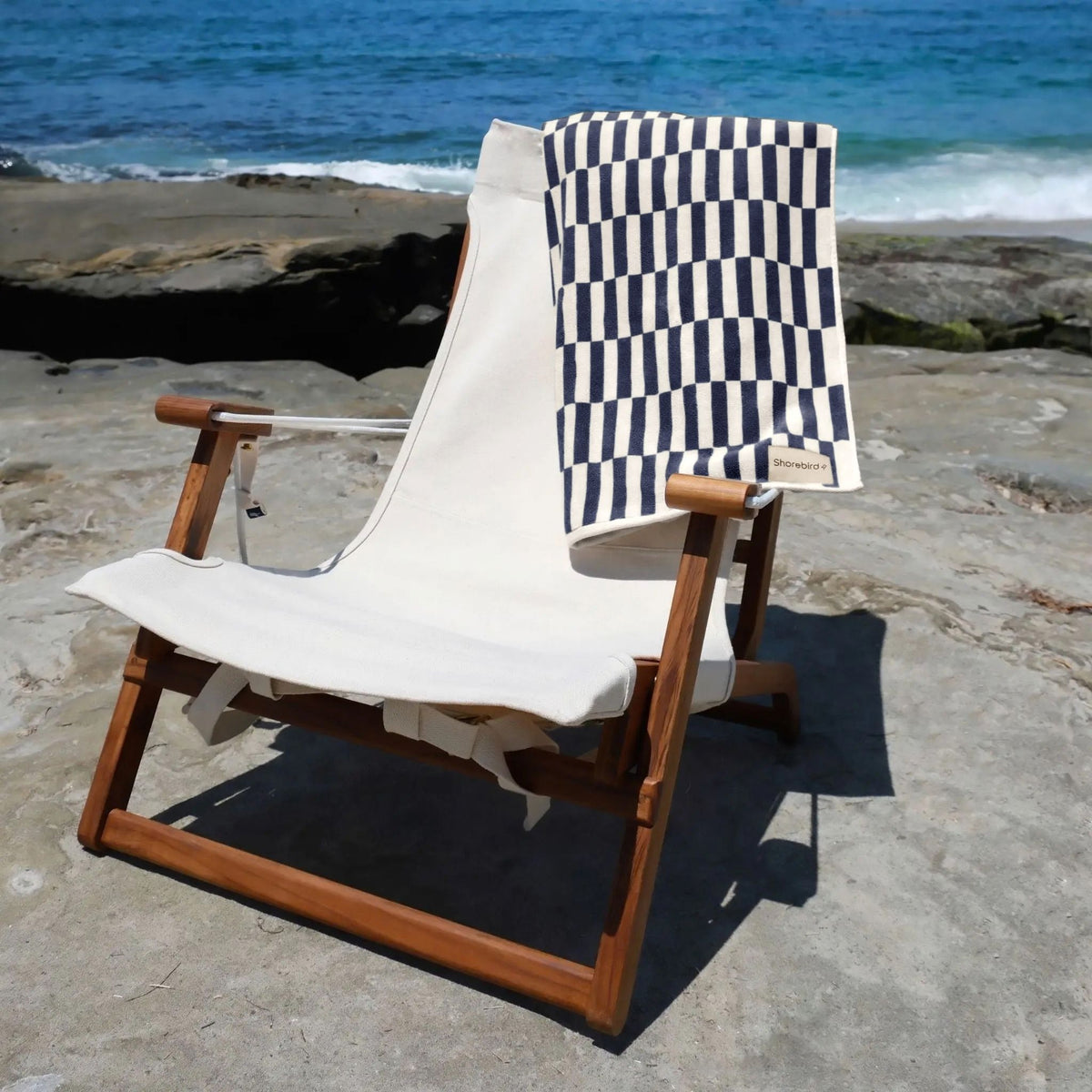 Navy check Oversized Beach Towel draped over the Shorebird teak beach chair on a rocky shore #color_nantucket