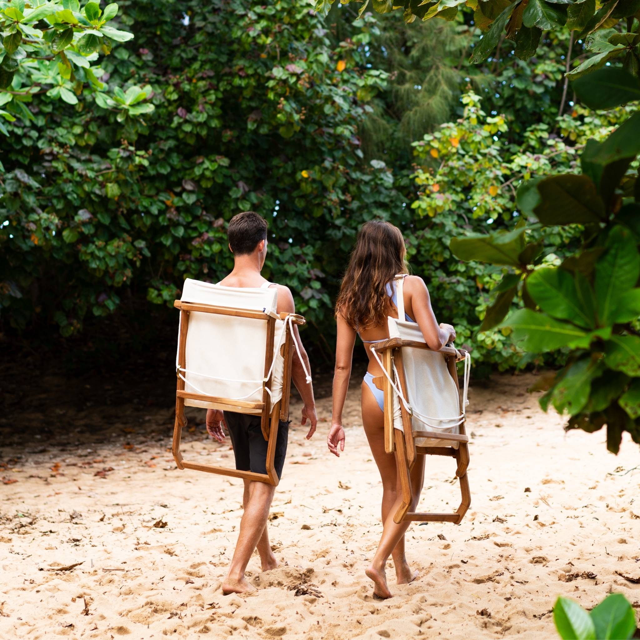 Two people walking on a sandy path with Shorebird Chairs on their backs, surrounded by greenery.